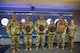 Six people dressed in Army green camouflage uniforms are posing for a picture while standing in front of a large screen monitor. Some people have wooden award plaques in their hands.