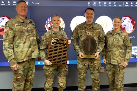 Three men and one woman dressed in Army green camouflage uniforms are posing for a picture while standing in front of a large screen monitor. The two middle people have wooden award plaques in their hands.