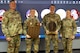 Three men and one woman dressed in Army green camouflage uniforms are posing for a picture while standing in front of a large screen monitor. The two middle people have wooden award plaques in their hands.