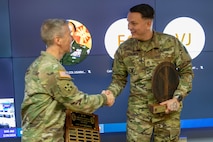 Two men dressed in Army green camouflage uniforms are shaking hands in front of a large monitor. They both have wooden plaques in their hands.