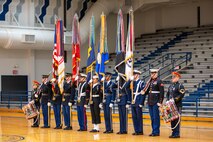 A color guard team is standing in a row holding flags, rifles and there is a drummer on each end. They are in a gymnasium with grey bleacher seats in the background.