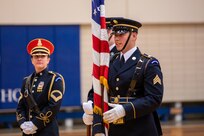 A man dressed in a dark Army ceremonial uniform with white gloves is talking while holding a US flag at his side. There is a woman dressed in a similar uniform with a round red hat on nearby in the backgroud. They are in a gymnasium.