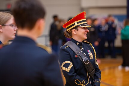 A woman dressed in a dark Army ceremonial uniform with a round red hat is smiling while looking away as she stands in a gymnasium with others looking on.