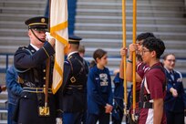 A man dressed in a dark Army ceremonial uniform is showing some students how to hold a ceremonial flag.