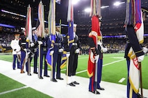 A color guard team dressed in various military uniforms is standing with large flags at their sides on the sideline of a football stadium with bright green turf.