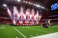 Fireworks are shooting into the air at 45-degree angles in a large stadium. There are people near the fireworks who are holding up a large US flag parallel with the ground. In the foreground on the right is a color guard team that is wearing various military uniforms while holding various flags.