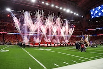 Fireworks are shooting into the air at 45-degree angles in a large stadium. There are people near the fireworks who are holding up a large US flag parallel with the ground. In the foreground on the right is a color guard team that is wearing various military uniforms while holding various flags.