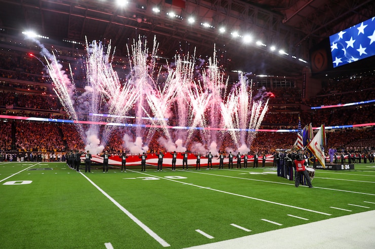 Fireworks are shooting into the air in a large stadium. There are people near the fireworks who are holding up a large US flag. On the right is a color guard team that is wearing various military uniforms while holding various flags.