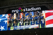 A color guard team who is holding various flags and wearing military uniforms is seen on the big screen of a large stadium. On the big screen are the letter NRG Stadium