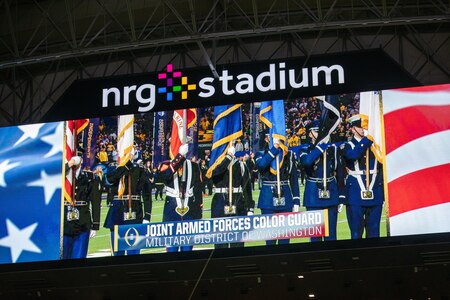 A color guard team who is holding various flags and wearing military uniforms is seen on the big screen of a large stadium. On the big screen are the letter NRG Stadium