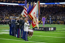 A color guard team dressed in various military uniforms is holding various flags with all of them lowered except the US flag in the middle. There is a drummer on each end and they are saluting.  In the distant background is a woman standind on a small black platform who is wearing a fluffy lavender top and lavender-colored leggings.