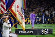 Some people in uniforms are seen on the far left holding various flags while a Black woman dressed in a furry lavender coat and pants is standing on a small black platform in the middle of a football field with bright green turf.