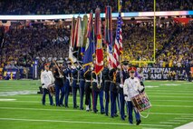Members from various branches of the military are carrying flags and drums off a football field in a large stadium.