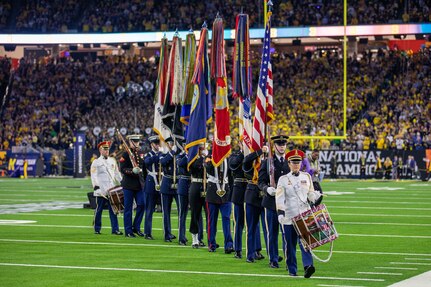 Members from various branches of the military are carrying flags and drums off a football field in a large stadium.
