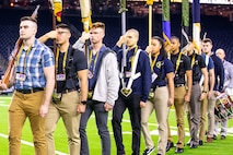 Several men and one woman are marching towards the camera and off of a footbal field with bright green turf. They are carrying a mix of rifles and flags, and are dressed in a variety of outfits in various colors.