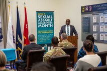 A Black man in a grey suit and striped tie is speaking behind a brown podium to several people who are seated and looking his way. Behind him are some flags and a tall banner that reads April is Sexual Assault Awareness and Prevention Month (SAAPM) and other illegible words.