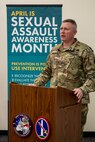 A man dressed in Army green camouflage is speaking from a brown pulpit. Behind him is a tall teal-colored sign with the words April is Sexual Assault Awareness and Prevention Month (SAAPM) and other illegible words