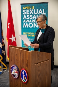A woman dressed in a dark suit and white blouse is holding a white piece of paper while reading at a brown podium. In the background is a teal-colored sign with words that read April is Sexual Assault Awareness and Prevention Month (SAAPM), among other illegible words. There is also a bright red flag with two white stars on it in the background