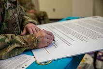 Someone's hand and forearm are resting on a table while they are writing on a large white poster. The person is wearing Army green camouflage. The table has a teal-colored tablecloth.