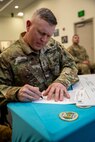 A man with silver and white short hair is writing on a white poster that is on a table with a teal-colored tablecloth. There is a round coin on the table in the foreground.  The man is dressed in Army green camouflage.