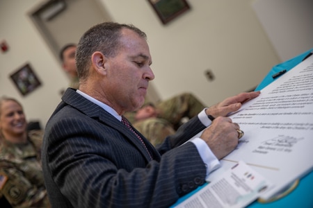 A white man with short darker hair wearing a dark grey black-striped suit is writing on a large white posterboard. There is someone smiling in the background who is wearing an Army green camouflage uniform.