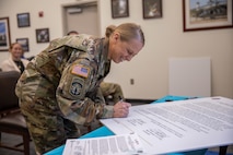 A female dressed in Army green camouflage uniform is smiling while leaning over a table signing a large poster. The table cloth under the poster is teal colored, and the woman's uniform has a red, white and blue US flag patch on the upper sleeve.