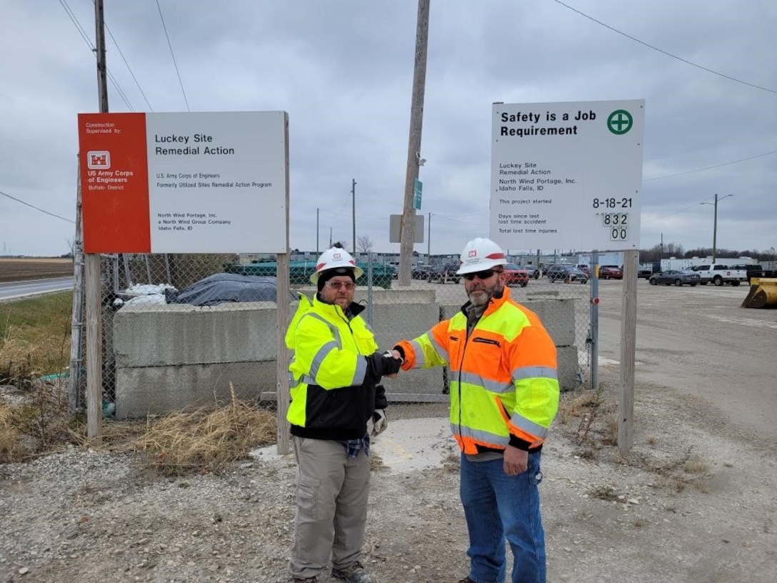 Two men in reflective coats and hard hats stand in front of construction site signs.