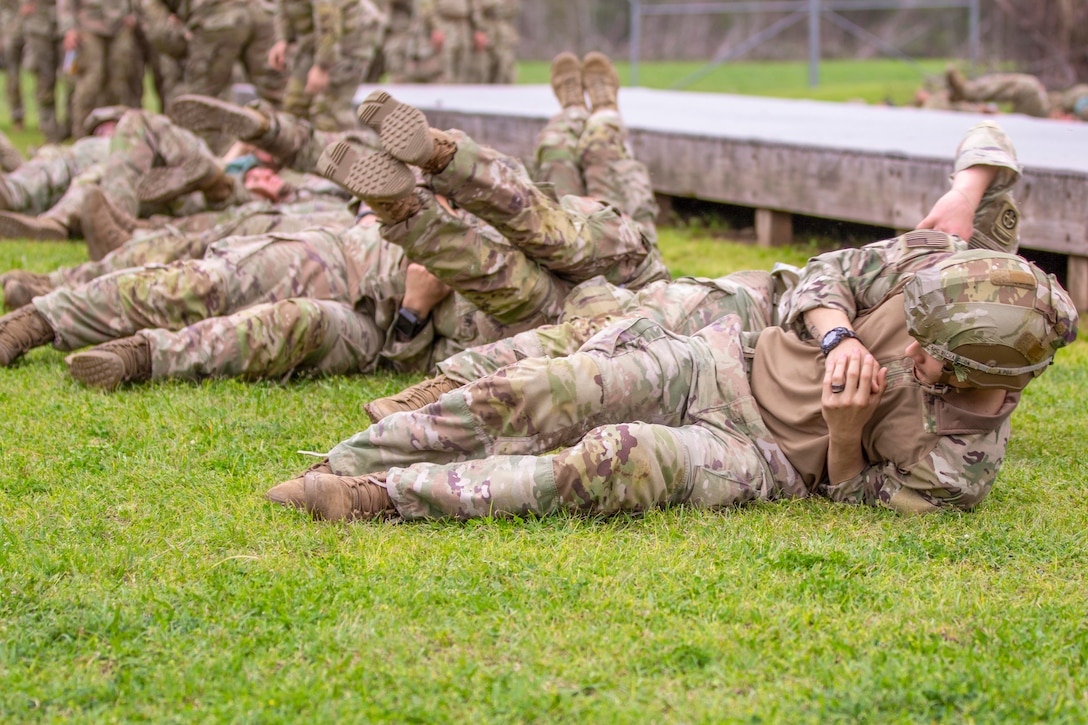 Soldiers roll on the grass in formation near a wooden structure as fellow soldiers stand in line in the background.