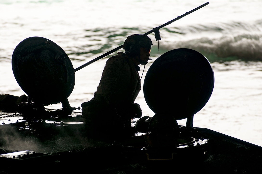 A Marine aboard a small boat stands near satellites as waves move in the background as seen in silhouette.