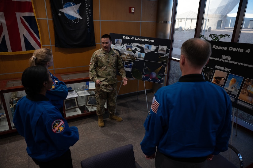 An airman stands in front of a poster in a conference room while three astronauts look on.