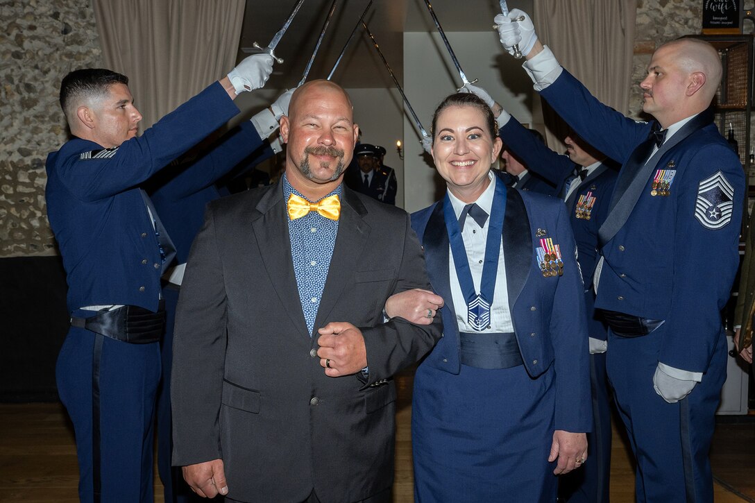 An airman walks arm-in-arm with a civilian in front of airmen holding up sabers in a room.