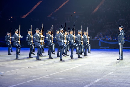 military personnel perform drill and ceremony movements before a large indoor crowd