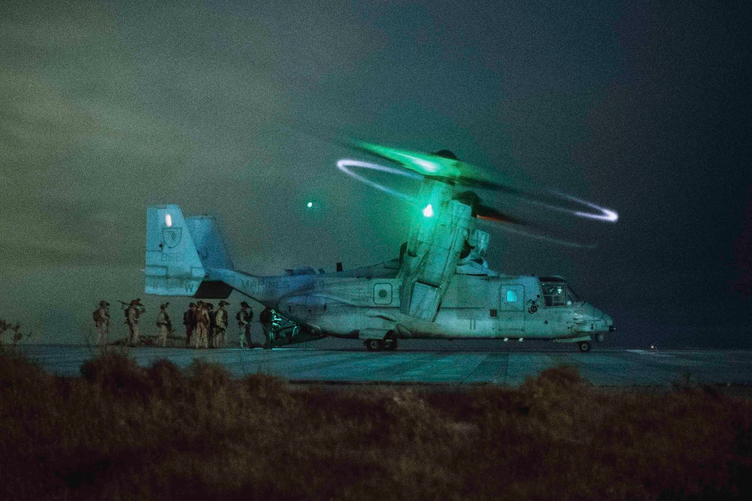 A group of Marines and sailors prepare to board a military aircraft under a greenish light at night.