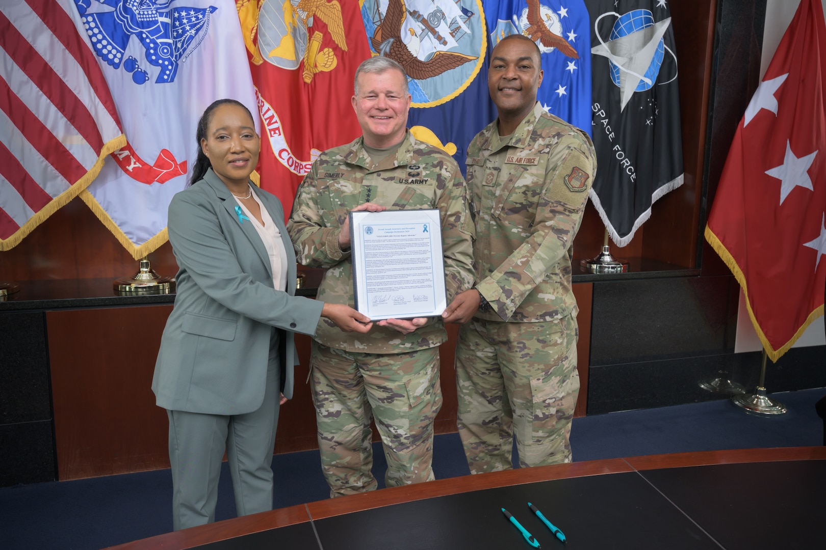 One woman and two men stand side by side in front of flags holding a piece of paper.