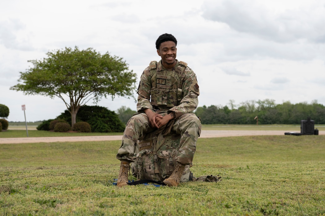 A photo of a person in battle rattle, sitting on his ruck, posing for a photo.