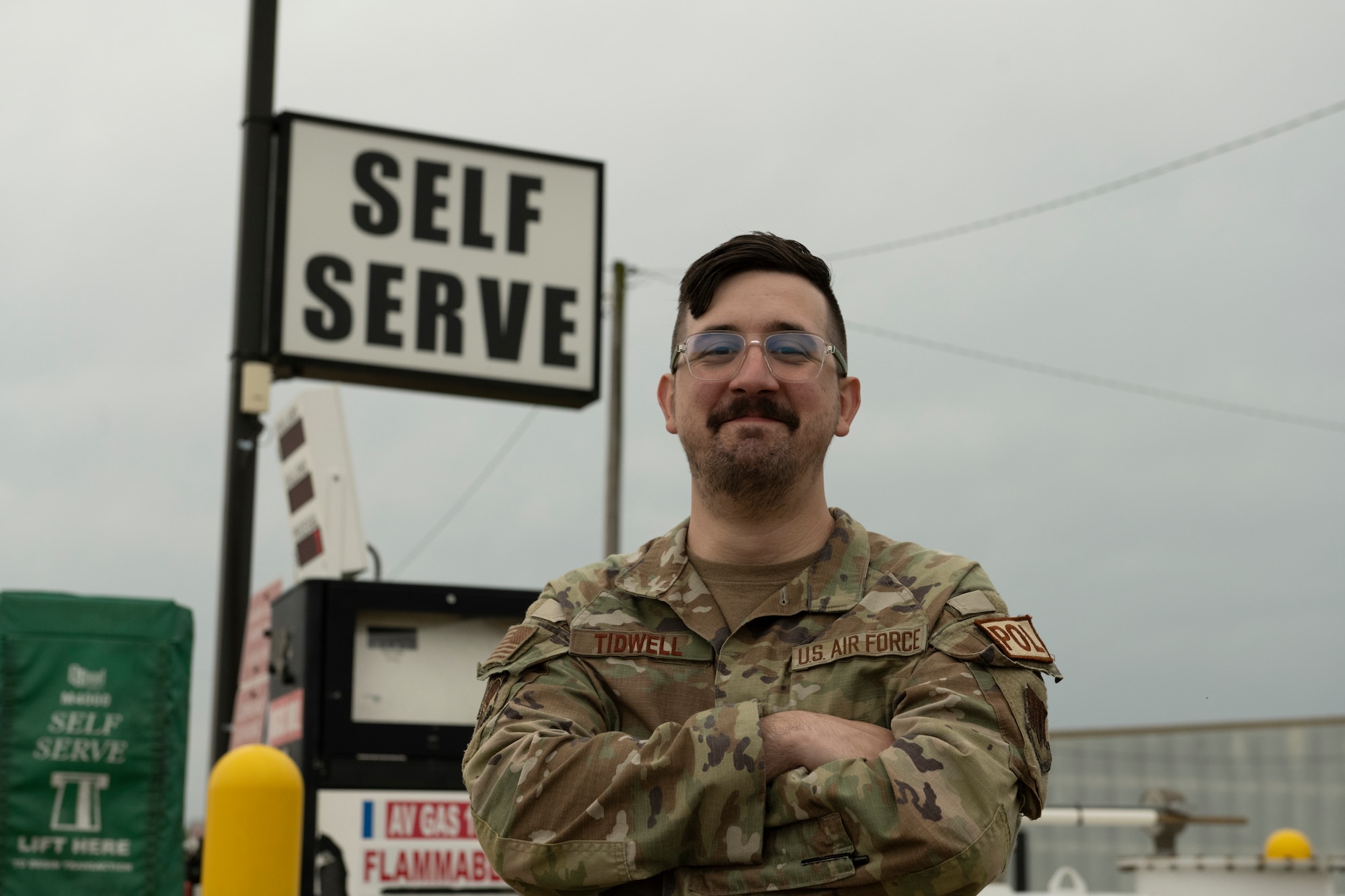 A photo of some one crossing their arms in front of them, with a gas station in the back ground.