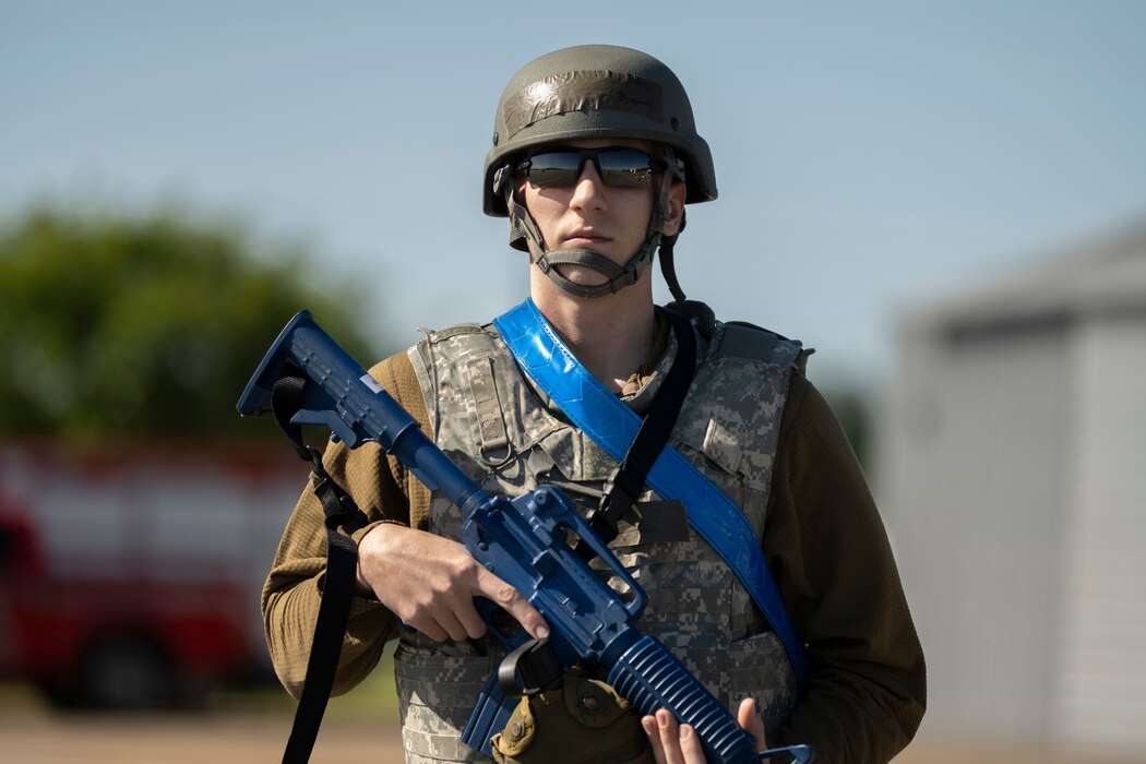 A photo of a person in battle rattle, holding a dummy gun, posing for a photo.