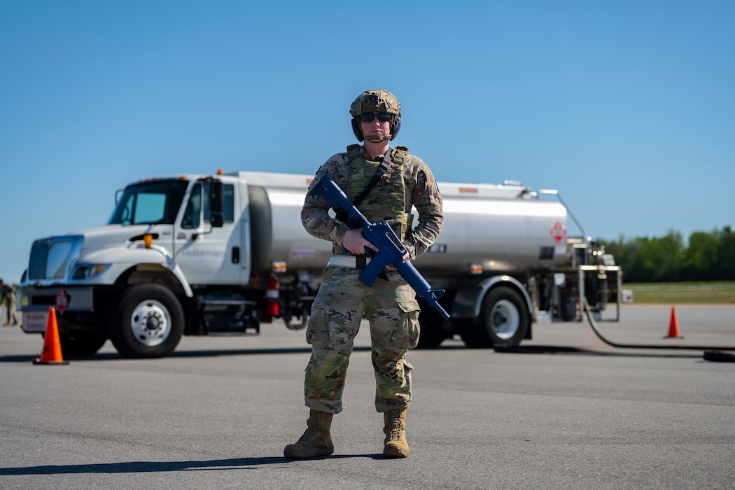 A photo of a person posing in battle rattle with a dummy gun, in front of a fuels truck.