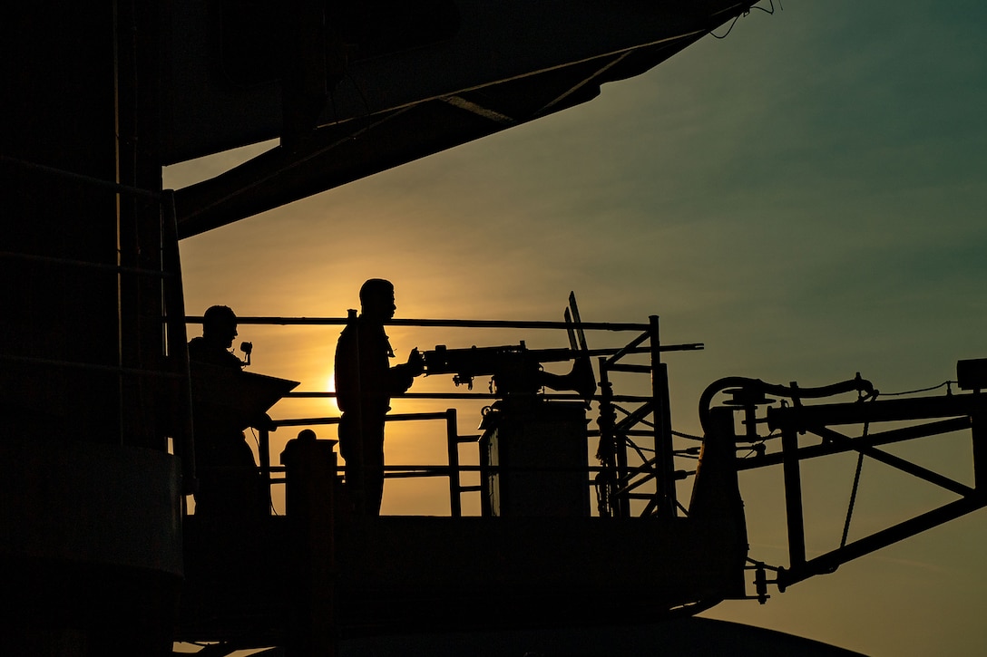 Two sailors stand watch at a mounted gun on a ship at twilight.