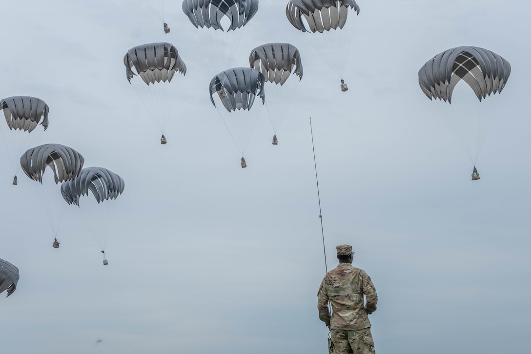 A soldier watches as military containers attached to parachutes fall from the sky.