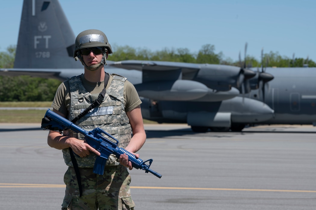 A person in battle rattle holding a dummy gun with an aircraft in the background.