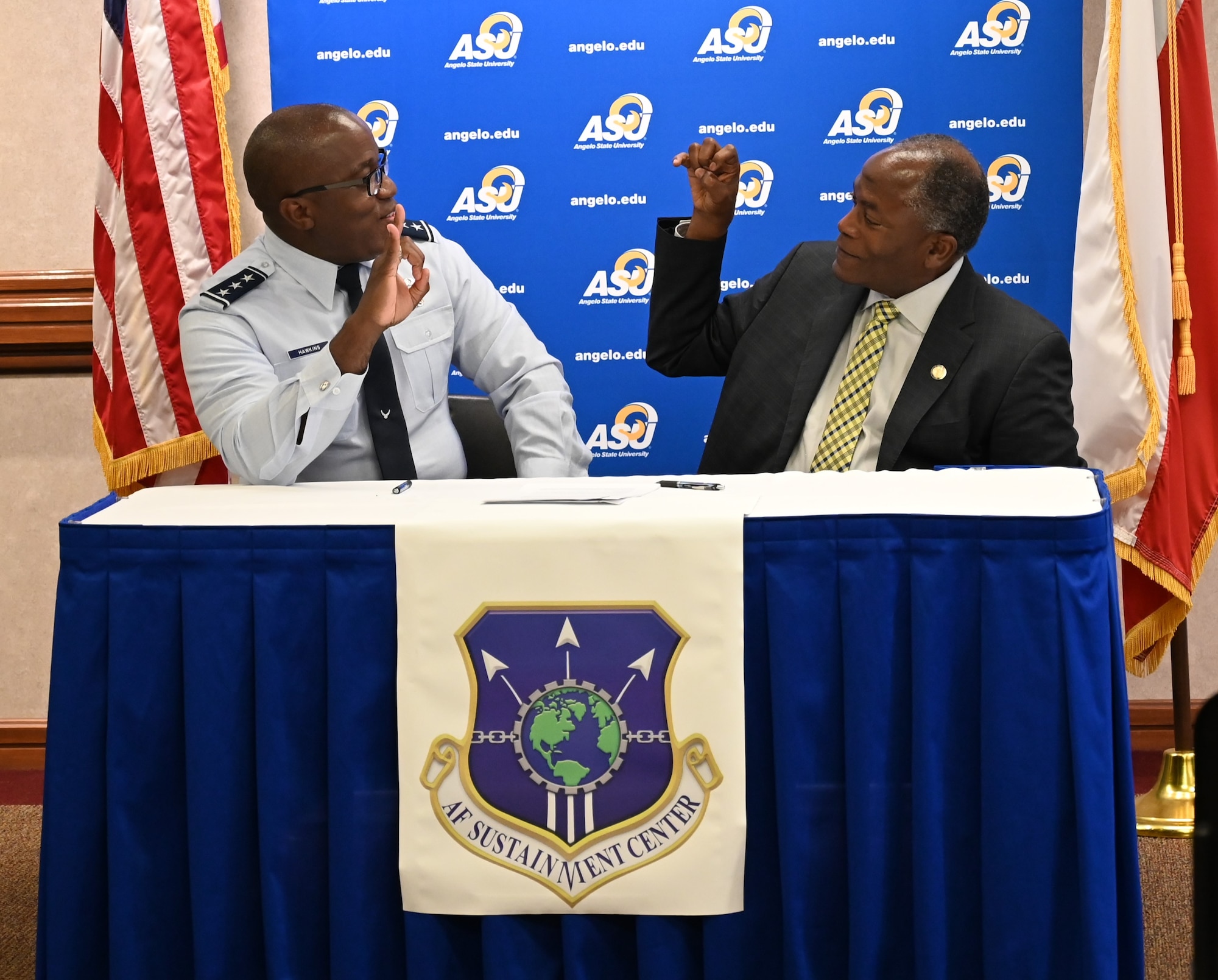 Two men sit at a table looking at each other while a making a sign with their right hands that represent the ASU Rams.