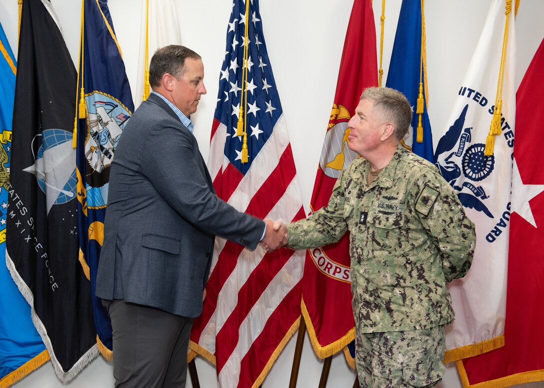 A light skinned man in a dark grey business suit shakes hands with a light skinned man in a Navy utility uniform.