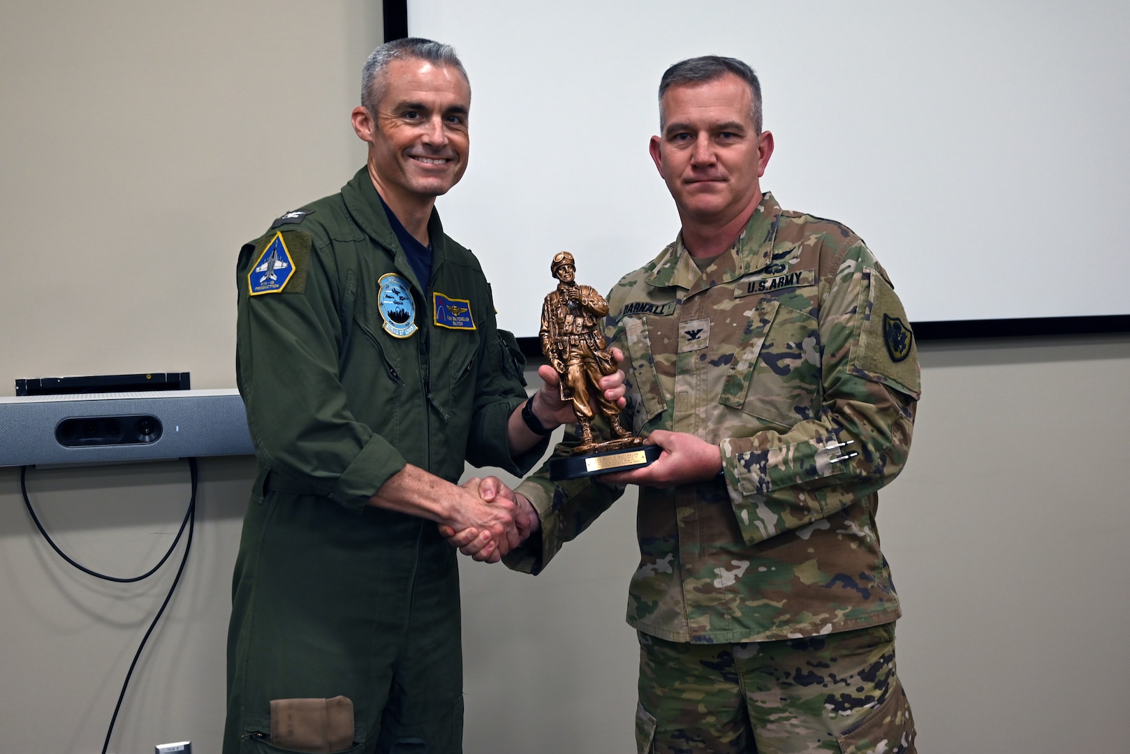 Man wearing a flight suit receives a statue from a man wearing a camouflage uniform