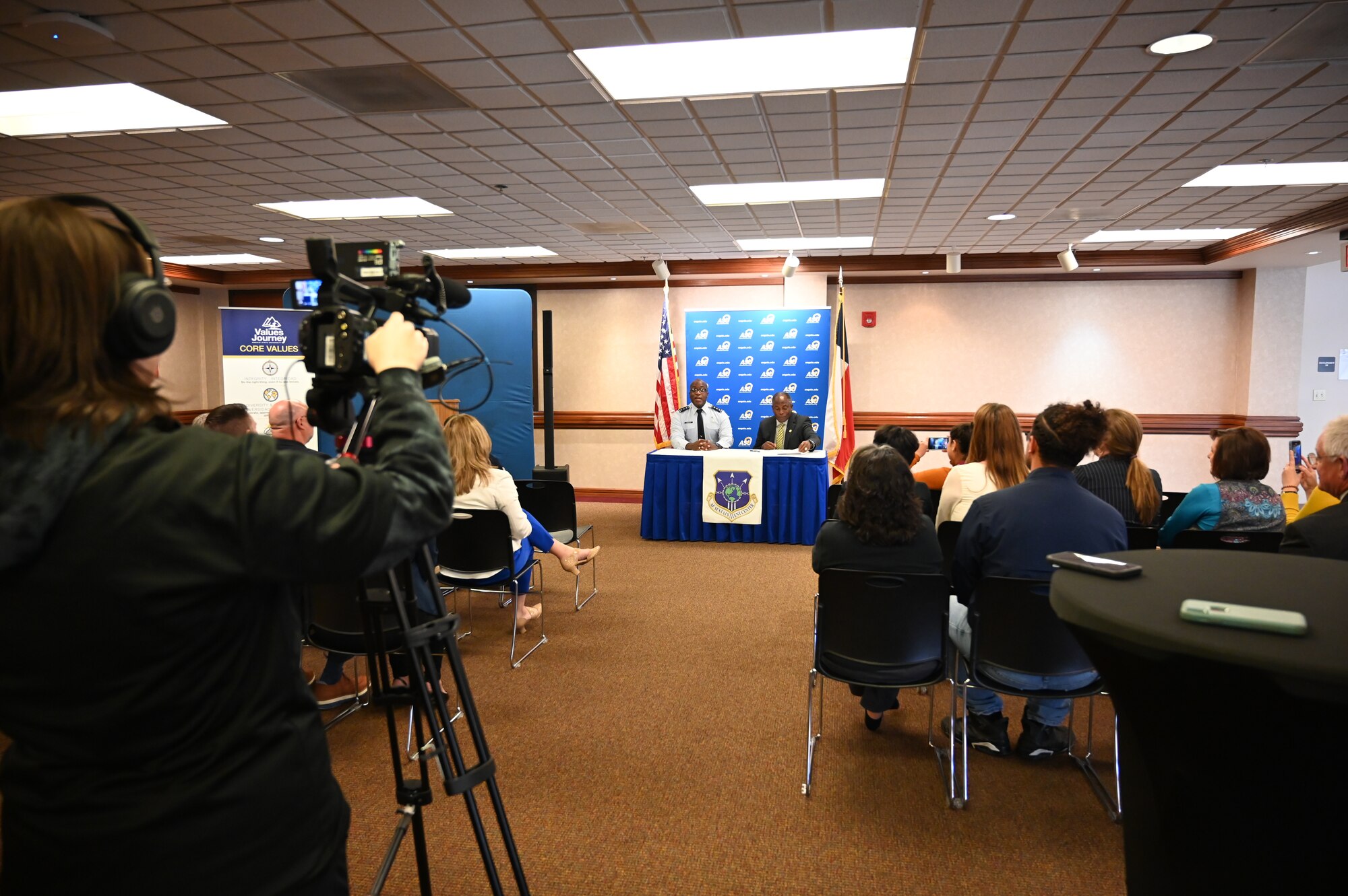 A wide shot showing two men signing papers at a table while a video camera records them and audience members look on.