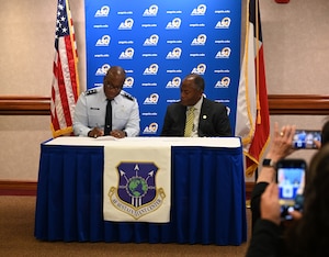 Two men sit at a table signing documents while the image is displayed on the cell phones of two audience members.