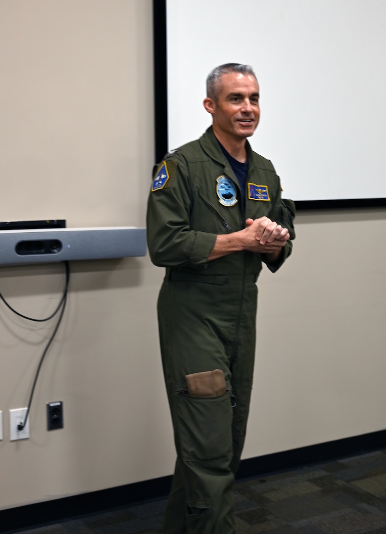 A smiling man wearing a flight suit is standing up and speaking