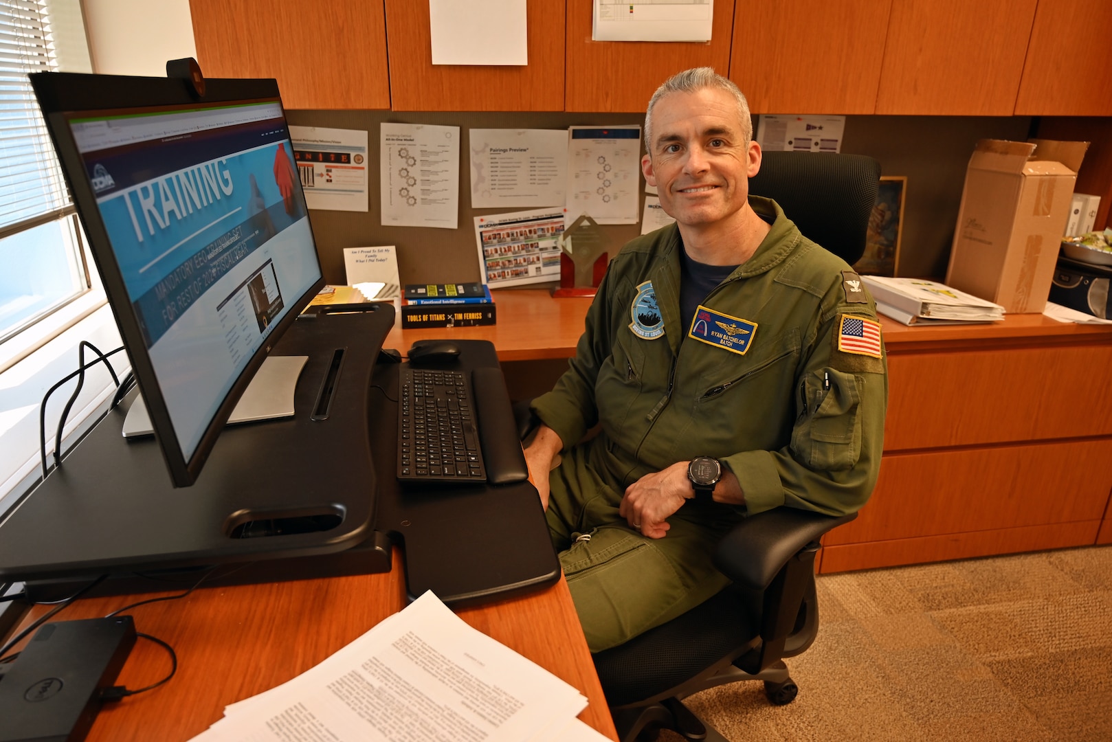 A smiling man wearing a flight suit sits at his desk near a computer