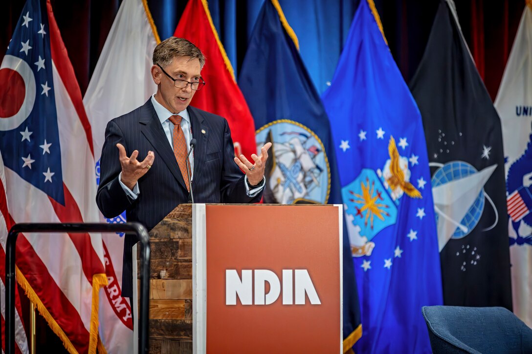 A man in a business suit gestures with his hands while speaking on stage in a ballroom.