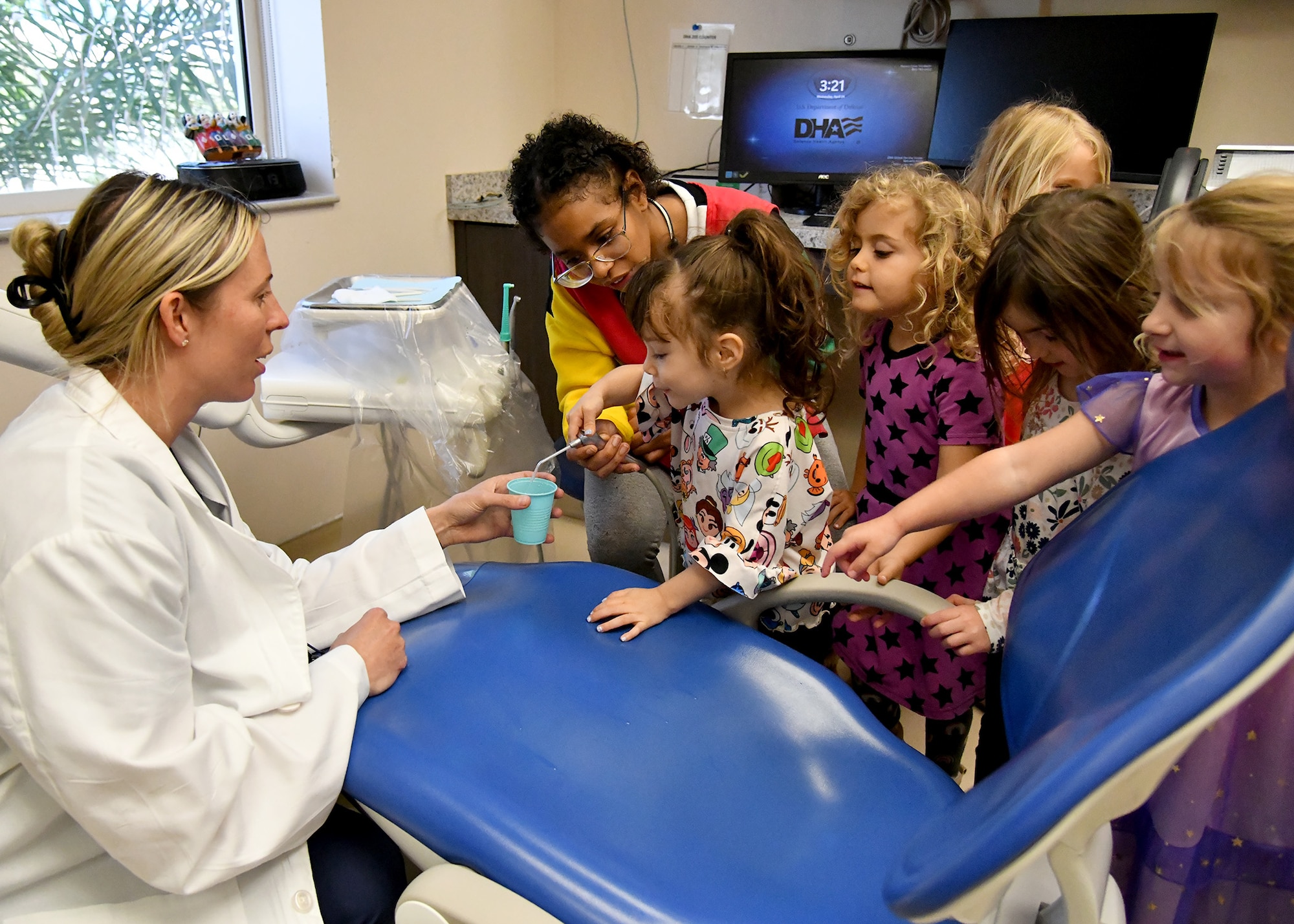 children gather around Dr. Baughman during their dental clinic visit to see how the suction works, sucking water from a paper cup.